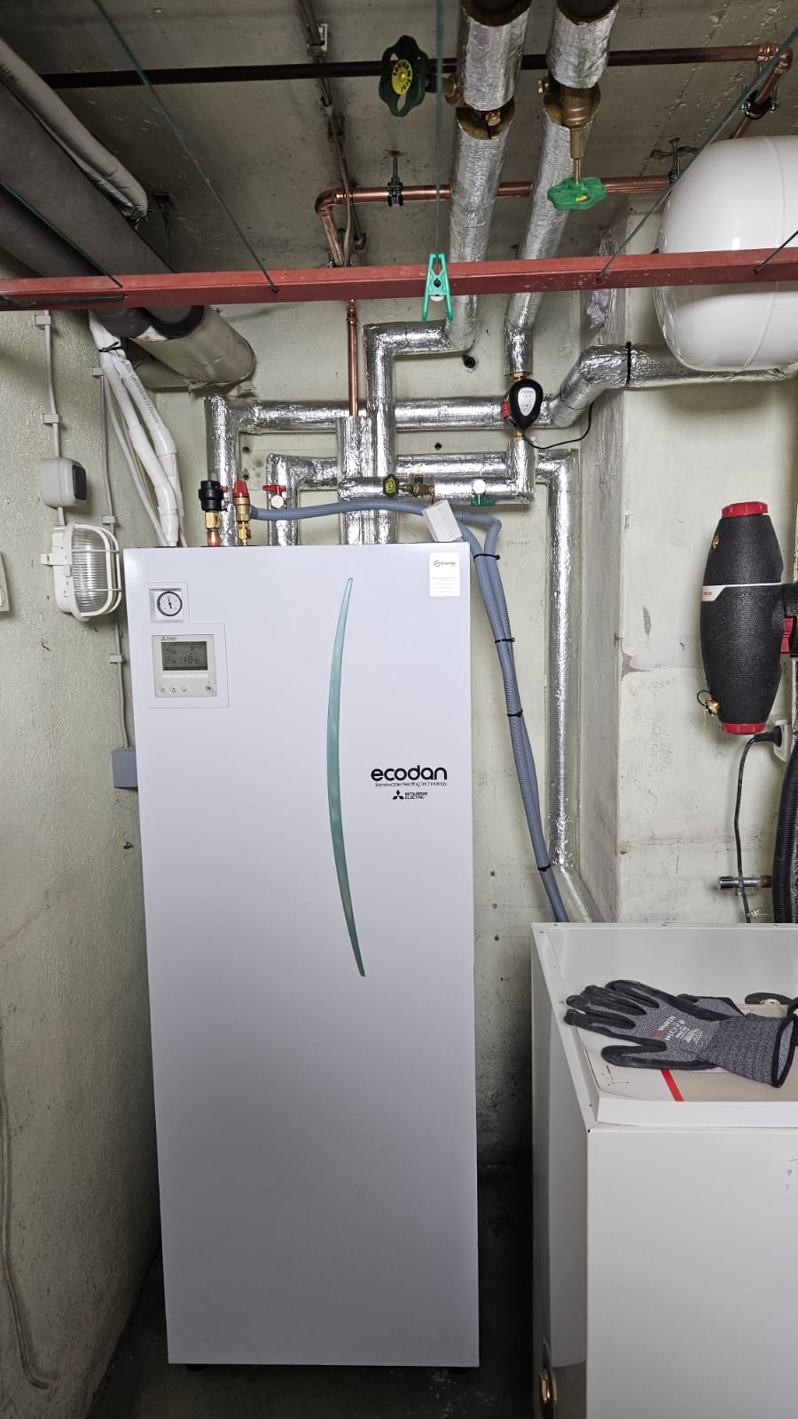 Heizungsanlage im Keller: Mitsubishi Ecodan Wärmepumpe mit angeschlossenen isolierten Leitungen und Steuerungseinheit.